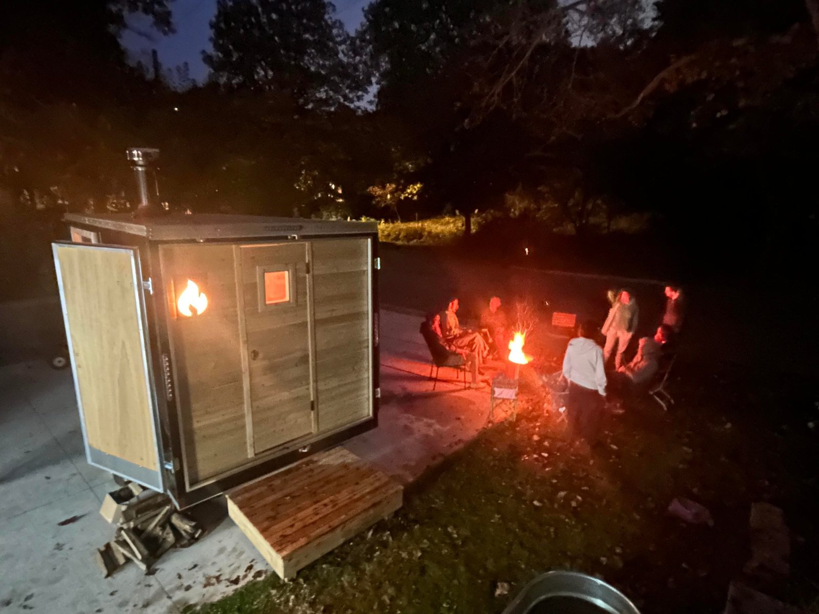 Night: group of people gathered around a firepit next to the glowing Thermic sauna.
