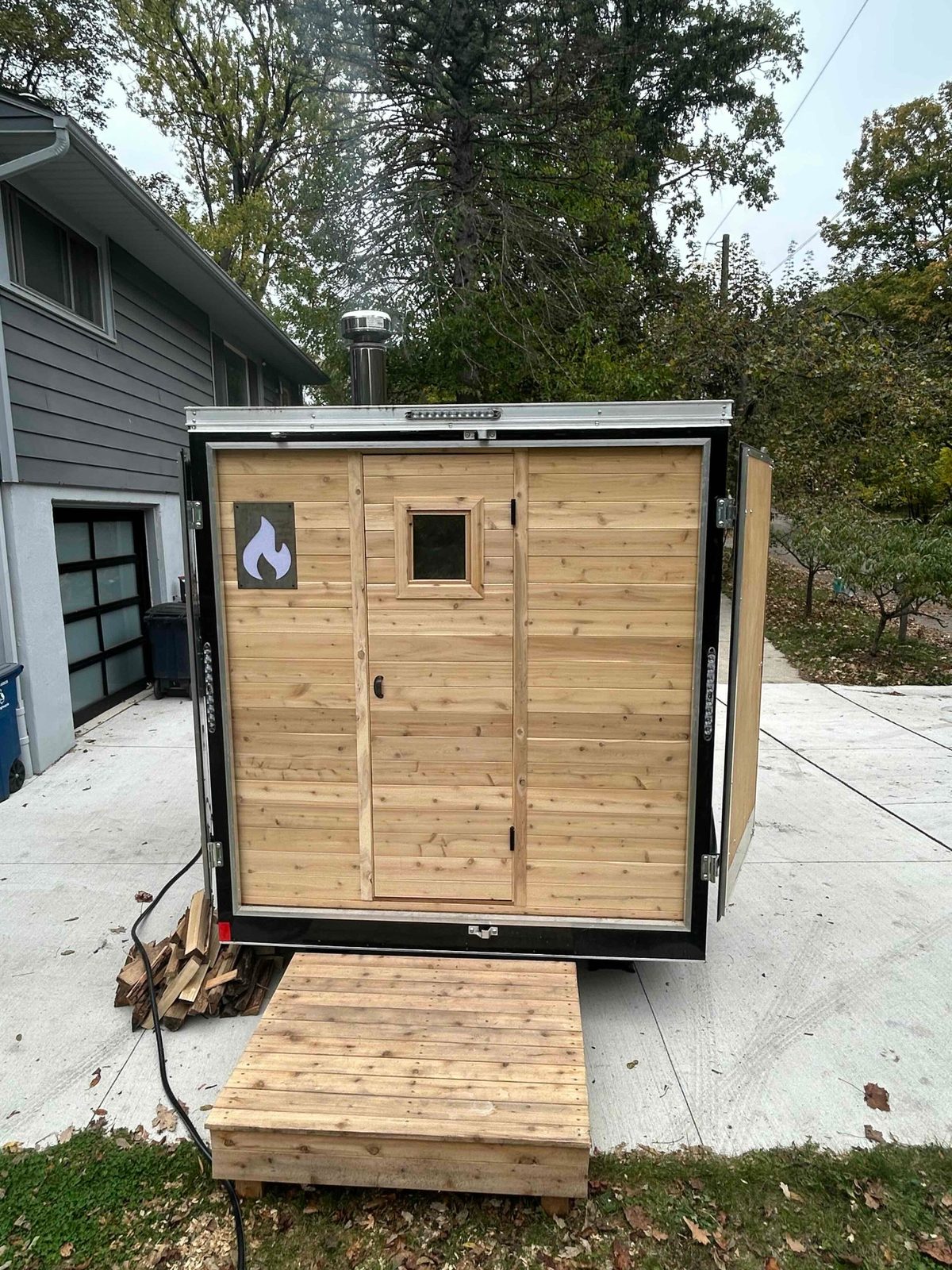 Back doors of the Thermic trailer open, revealing cedar-paneled sauna, cedar step, firewood stacked beside.